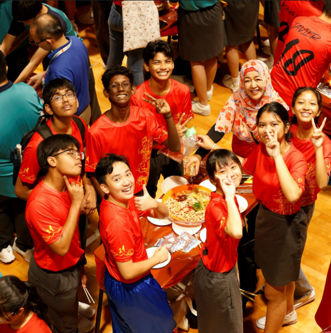 Overhead view of a large group of smiling children in red shirts gathered around a plate of food on a wooden floor.